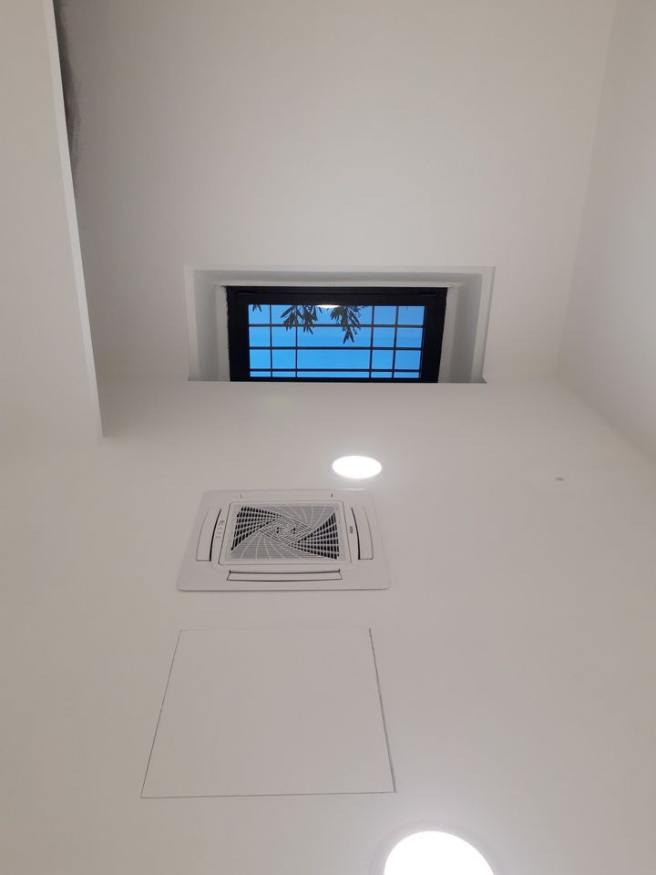 Controsoffitto bianco con faretti, unità di climatizzazione e botola d'ispezione, accanto a una finestra alta